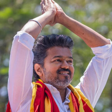 Man outdoors with arms raised in a prayer-like gesture, wearing a white shirt and a red-yellow scarf, smile on his face.