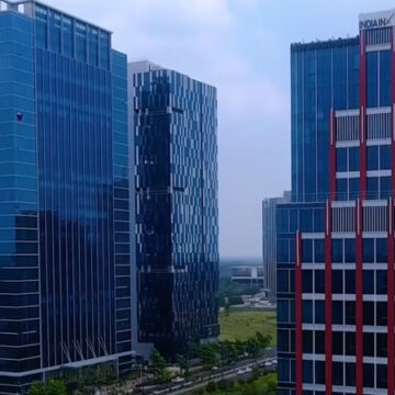 Modern blue glass office towers in a business district, with a road and green space below.