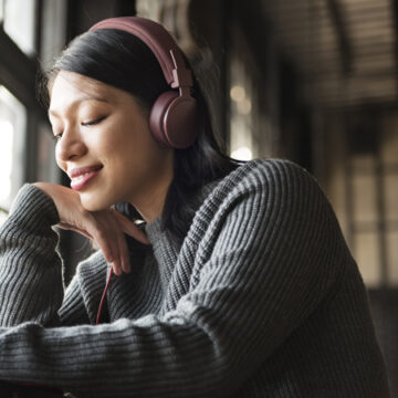 Young woman in a gray knit sweater with pink headphones smiles as she writes at a desk by a sunlit window.